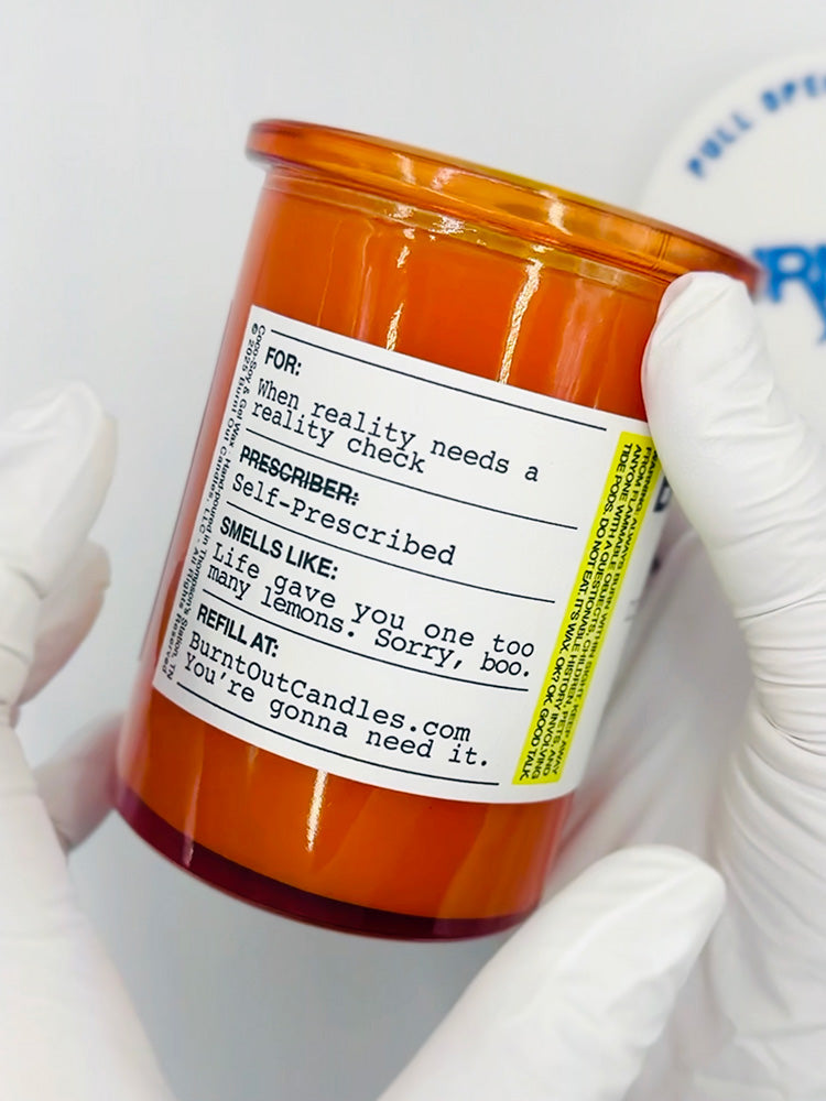 Orange candle jar with a label held by white gloved hands against a white background