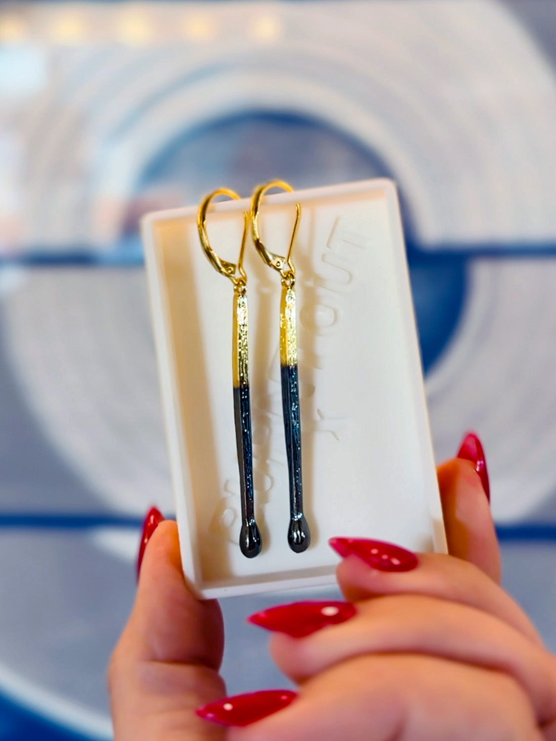 "burnt matchstick" gold earrings in a custom matchbox case held by a person with red nails.