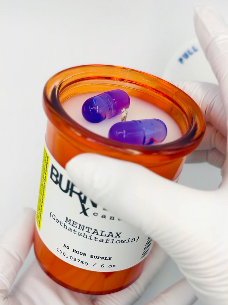 Candle in a bottle labeled 'Mentalax' with purple wax "pills" inside, held by a gloved hand.
