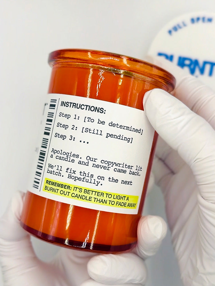 Orange candle with a humorous label held by a gloved hand against a white background.